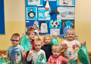 Na pierwszym planie stoi grupa dzieci i trzyma nagrody w zielonych reklamówkach. Z tyłu na tablicy umieszczone są prace plastyczne wykonane na konkurs. Prace przedstawiają zimowe zwierzęta.